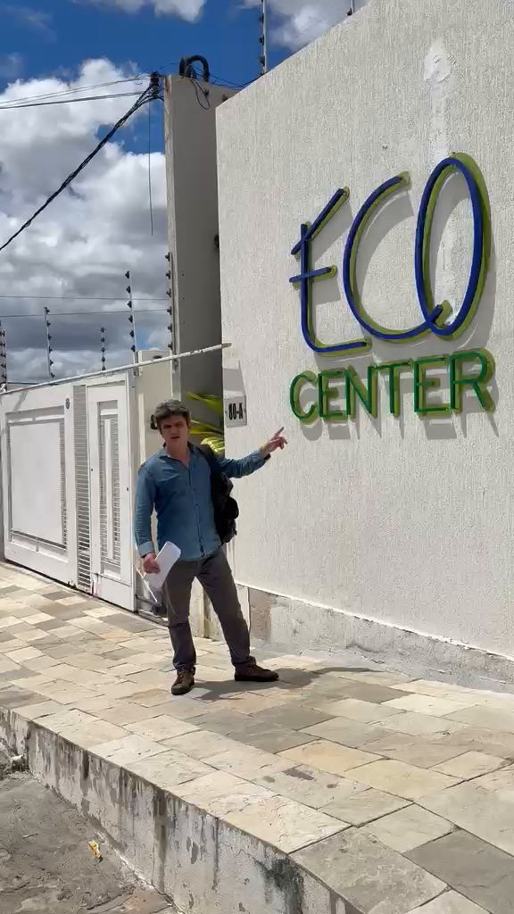 Angelo Girotto segura um documento e aponta para a fachada ECO CENTER; a numeração 80-A aparece ao lado do portão.