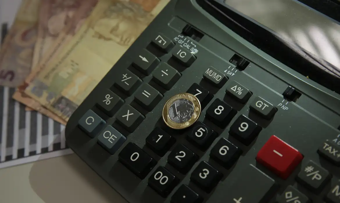 Moeda de um real sobre uma calculadora, com cédulas ao fundo.