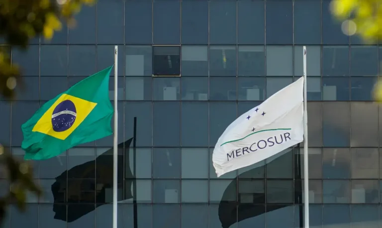 Bandeiras do Brasil e do Mercosul hasteadas lado a lado diante de um prédio envidraçado em Brasília.