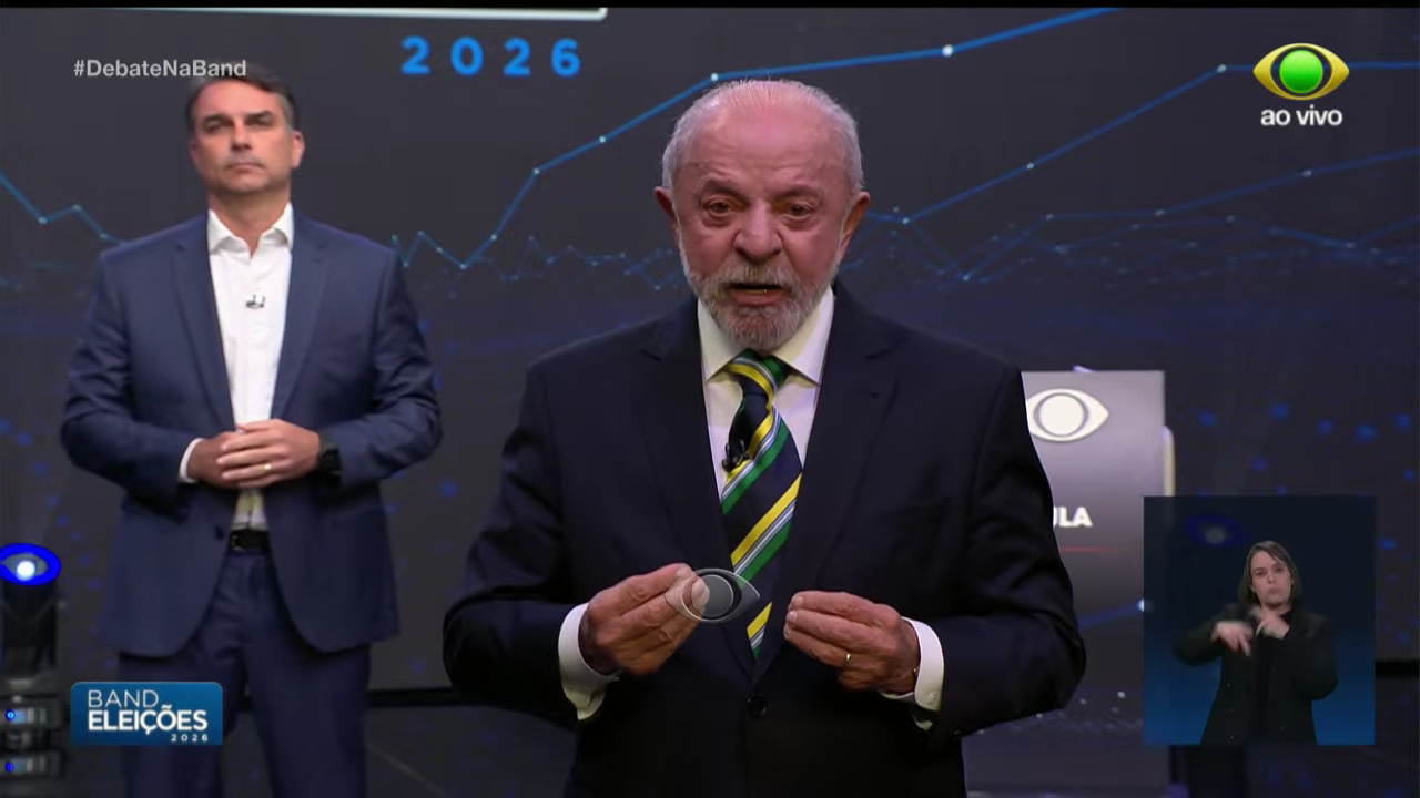 Lula fala em primeiro plano e Flávio Bolsonaro aparece ao fundo no palco do debate da Band.